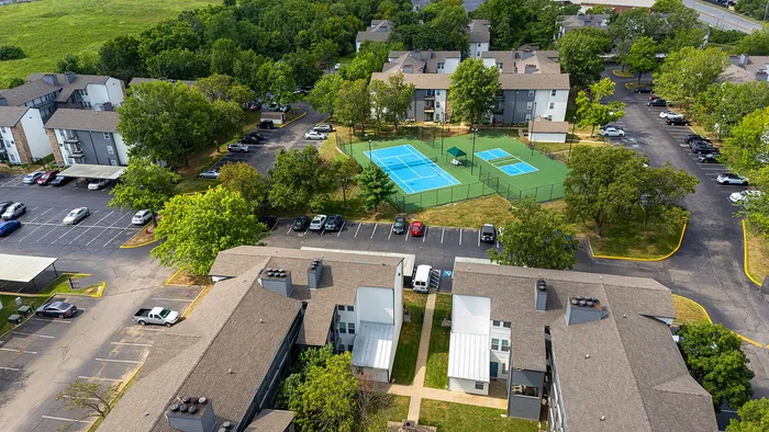 Bird's eye view of a modern apartment complex featuring lush greenery, tennis courts, and ample parking, offering a vibrant community lifestyle.
