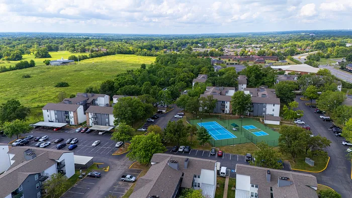 Aerial view of a serene apartment complex surrounded by lush greenery, featuring ample parking and vibrant tennis courts.