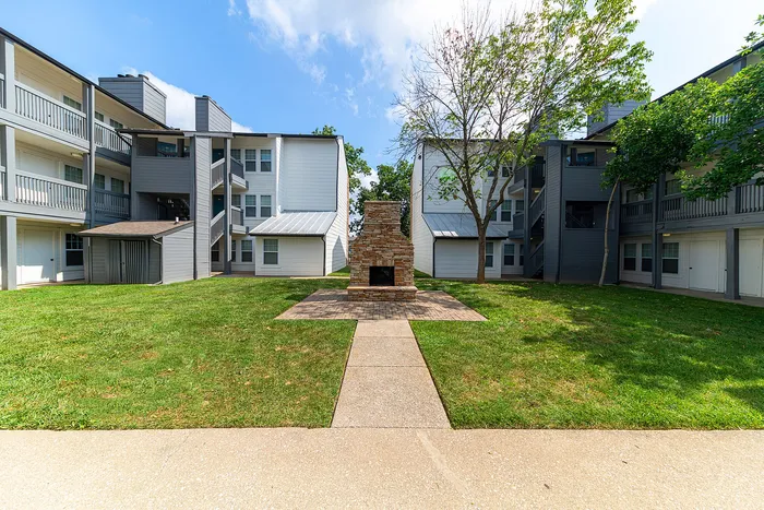 Charming apartment complex exterior featuring modern architecture with a central stone fireplace, surrounded by lush green lawns and trees.