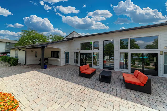 Vibrant outdoor patio area with modern seating and large glass doors under a bright blue sky with fluffy clouds.
