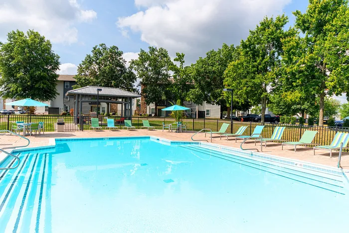 Inviting apartment complex pool with lush greenery, sun loungers, and shaded seating areas under a clear blue sky.