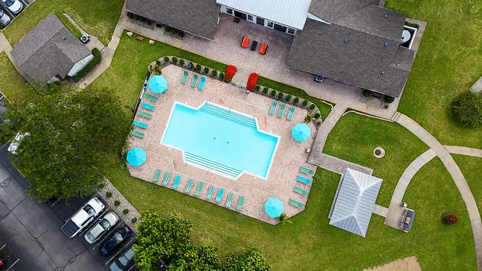 Charming aerial view of a refreshing pool with blue umbrellas and sun loungers, surrounded by lush green grass and cozy residential buildings.