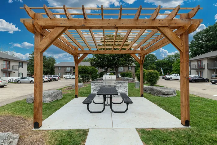Charming outdoor space featuring a wooden pergola with picnic tables and a barbecue grill, perfect for social gatherings in a serene apartment community.