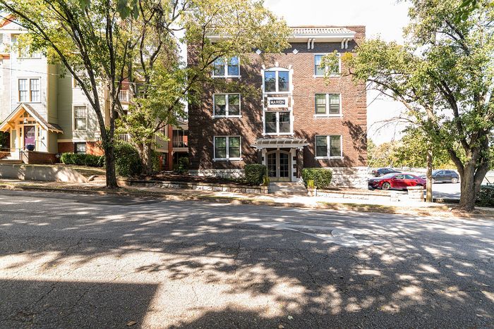 Charming brick apartment building surrounded by lush trees, offering a welcoming entrance and convenient parking.