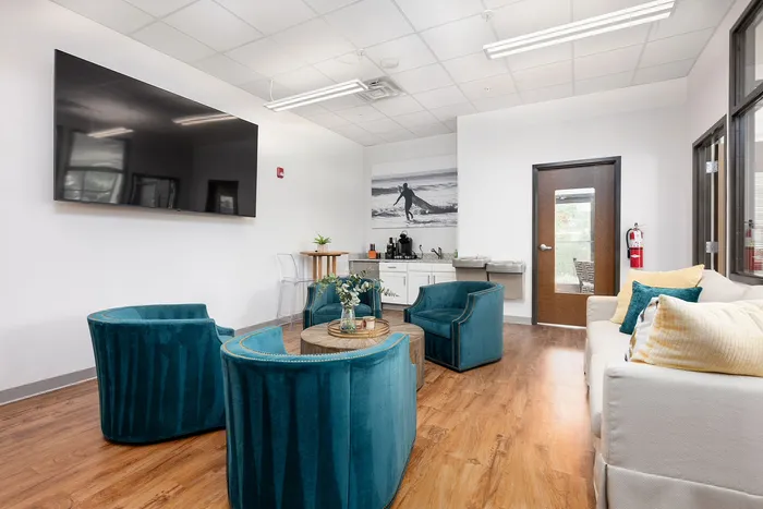 Chic lounge area with modern teal armchairs, a cozy sofa, and sleek wooden flooring, enhanced by natural light and a large wall-mounted TV.