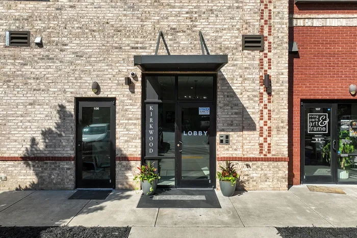 Charming brick exterior of Kirkwood building with a welcoming lobby entrance, flanked by modern planters, under bright sunlight.