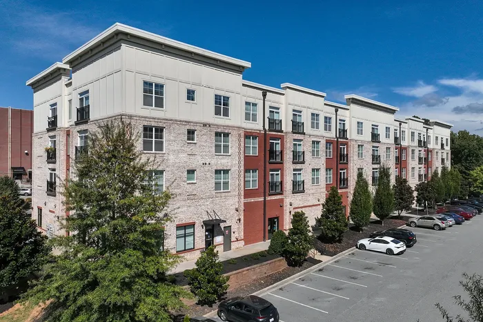 Modern four-story apartment building with a mix of red brick and cream-colored facade, surrounded by lush greenery and ample parking.