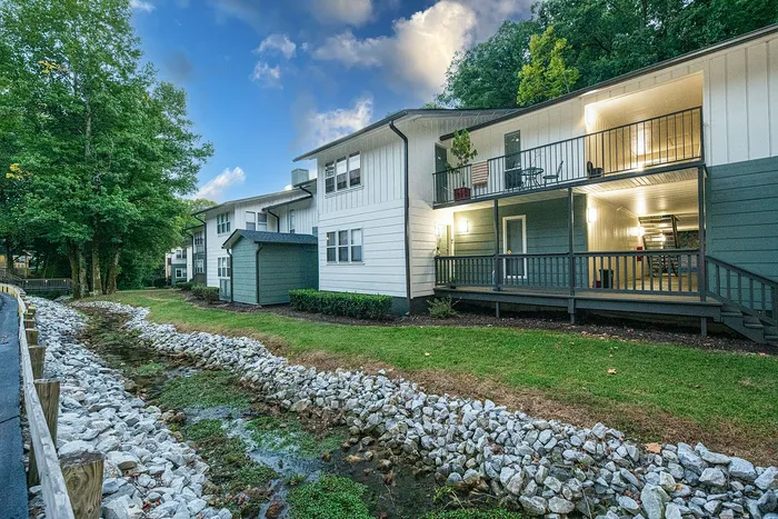 Charming two-story apartment building with spacious balconies and lush green surroundings, featuring a tranquil river rock-lined stream and scenic wooded backdrop.