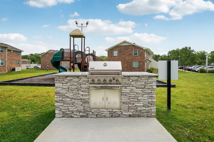 Inviting outdoor community space featuring a modern stone barbecue setup with a playful children's playground surrounded by lush green lawns and charming brick buildings.
