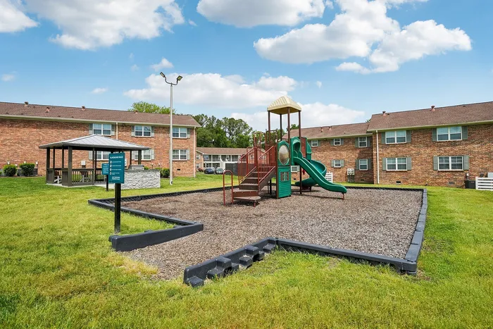 Charming apartment complex with a well-maintained playground, featuring a colorful slide and climbing structure, nestled in a lush green lawn under a clear blue sky.