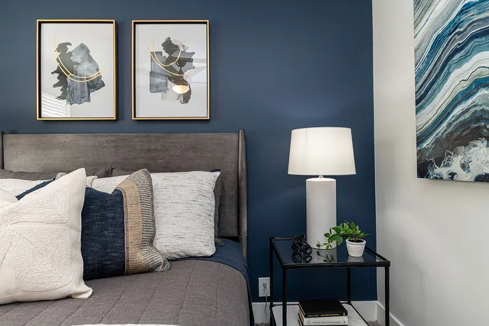 Elegant bedroom with plush bedding, modern art on deep blue walls, and a sleek lamp on a minimalist side table.
