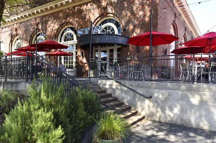 Charming brick building with vibrant red umbrellas, inviting outdoor dining area, and lush greenery on a sunny day.