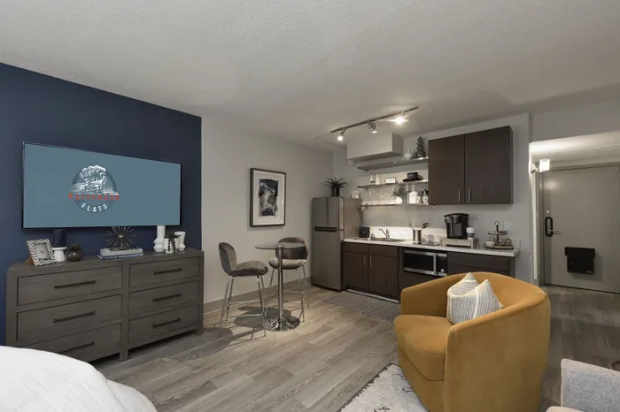 Modern studio apartment with a stylish kitchenette, cozy seating area, and a sleek wall-mounted TV. Elegant wood flooring complements the chic decor.