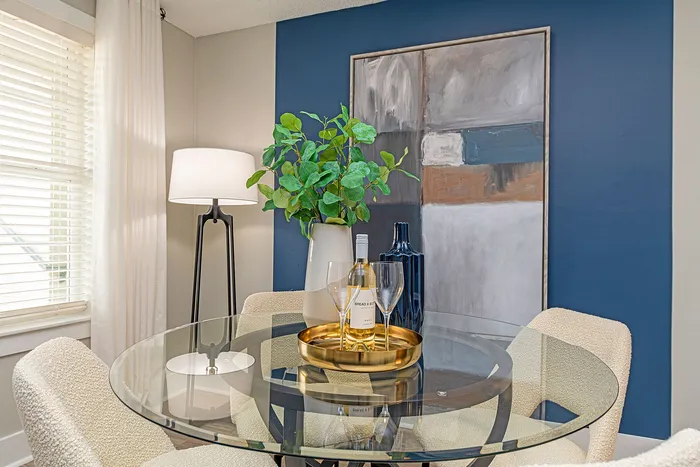 Elegant dining nook featuring a round glass table, plush chairs, and modern decor with a vibrant green plant and abstract artwork.