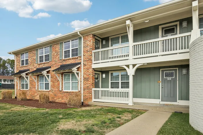 Charming two-story brick apartment building with classic white trim, featuring welcoming balconies and vibrant green lawn under a bright blue sky.