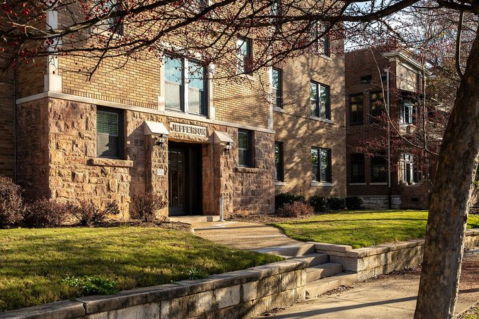 Charming apartment building with classic brick facade surrounded by lush greenery and shaded pathway.