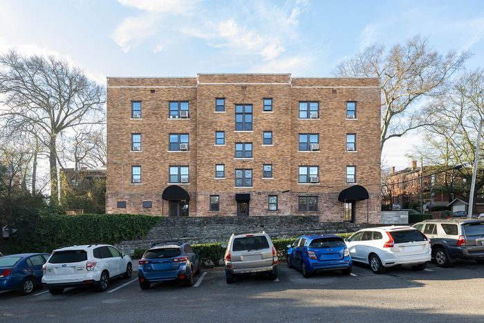 Charming brick apartment building with a clean, well-maintained facade, ample parking, and surrounded by lush greenery.