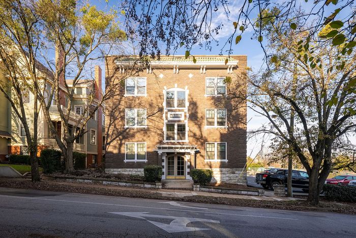 Charming brick apartment building with classic architecture, surrounded by mature trees on a sunny street.