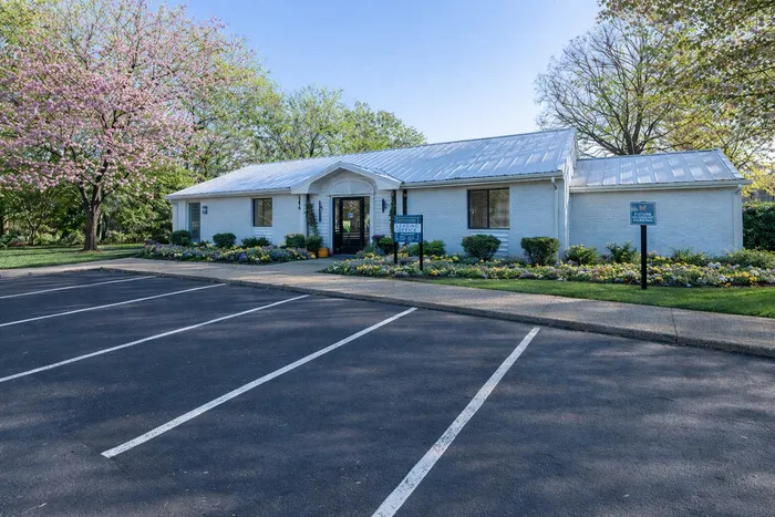 Charming white brick building with a modern metal roof, surrounded by lush landscaping and colorful flower beds, set against a backdrop of blooming trees and a spacious parking area.