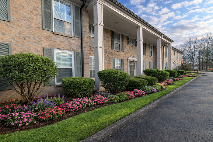 Charming brick apartment building with elegant columns and vibrant landscaped gardens under a blue sky.