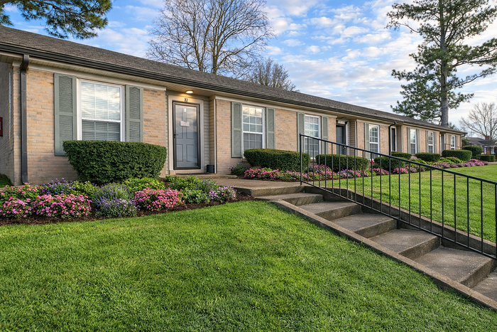 Charming brick apartment with inviting front landscaping, featuring vibrant flowers and a well-maintained lawn, under a clear blue sky.