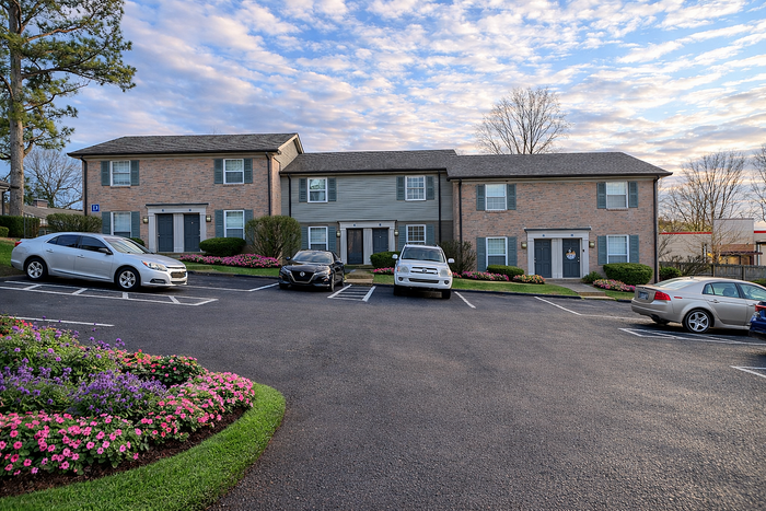 Charming apartment building with brick facade, surrounded by vibrant flower beds and ample parking under a picturesque sky.