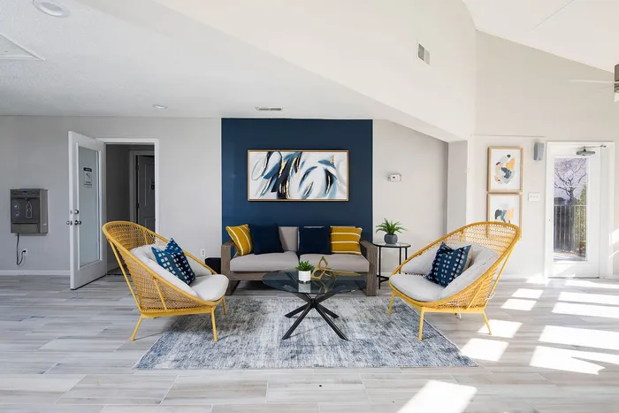 Chic living room with modern decor, featuring cozy wicker chairs, a stylish gray sofa, a glass coffee table, and vibrant artwork against a deep blue accent wall.