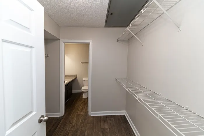 Spacious walk-in closet with sleek white shelving and a view into a modern bathroom featuring elegant fixtures and wood-style flooring.