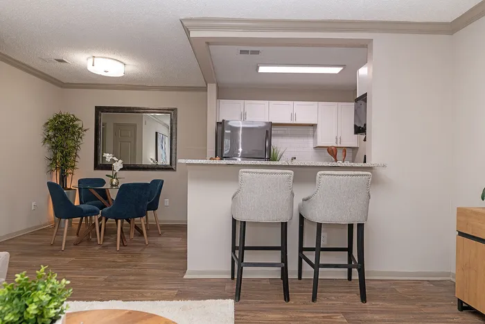 Modern apartment interior featuring a sleek kitchen with granite countertops, stylish breakfast bar with chairs, and cozy dining area with blue velvet chairs and a decorative mirror.