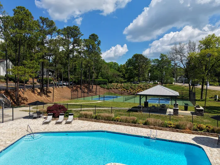 Inviting outdoor recreation area featuring a glistening pool, sun loungers, and tennis courts surrounded by lush greenery and tall trees.