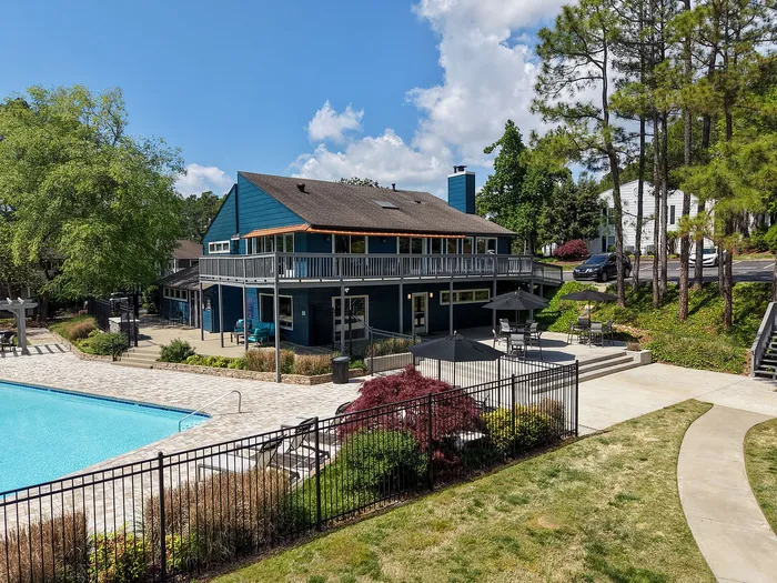 Charming apartment complex showcasing a vibrant clubhouse with poolside seating, surrounded by lush greenery and a sparkling blue swimming pool under a clear sky.