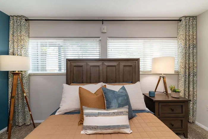 Cozy bedroom with elegant wooden headboard and chic accent pillows, featuring soft lighting from stylish floor lamps and natural light through large windows.