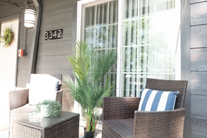 Charming apartment entrance with cozy wicker chairs, vibrant green plants, and elegant sliding glass door, creating a welcoming outdoor space.