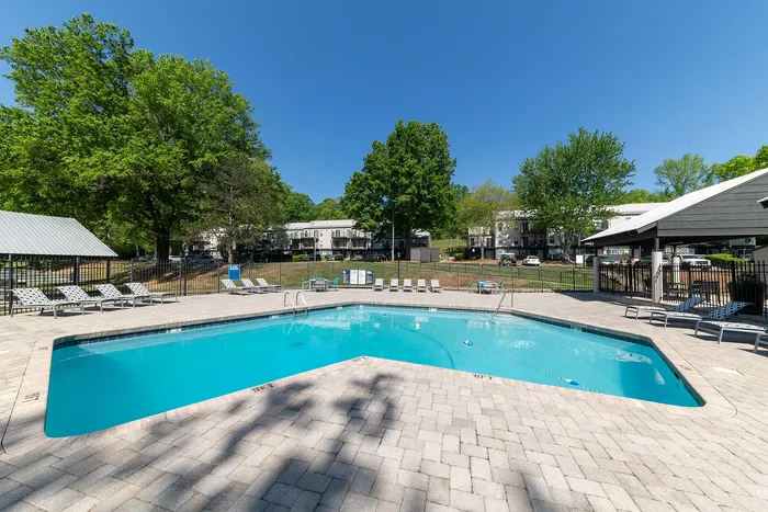 Inviting outdoor pool area with comfortable lounge chairs, surrounded by lush green trees and modern apartment buildings under a clear blue sky.