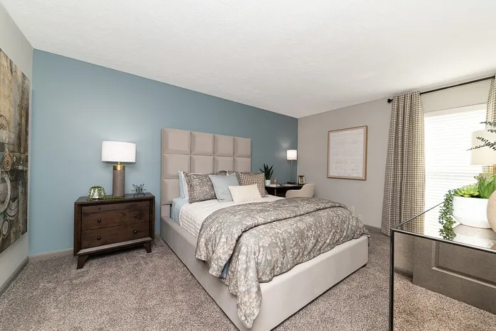 Inviting bedroom with plush bedding, stylish decor, and serene blue accent wall next to a sunlit window.