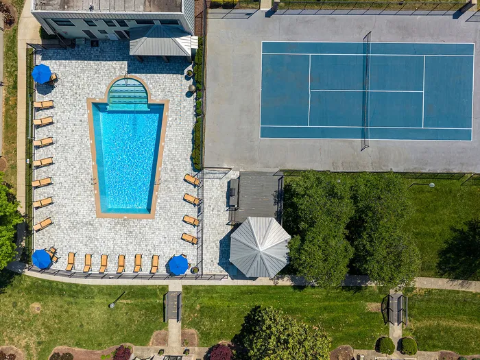 Aerial view of luxury apartment amenities featuring a sparkling blue swimming pool with sun loungers and umbrellas, adjacent to a well-maintained tennis court, set amidst lush green landscaping.