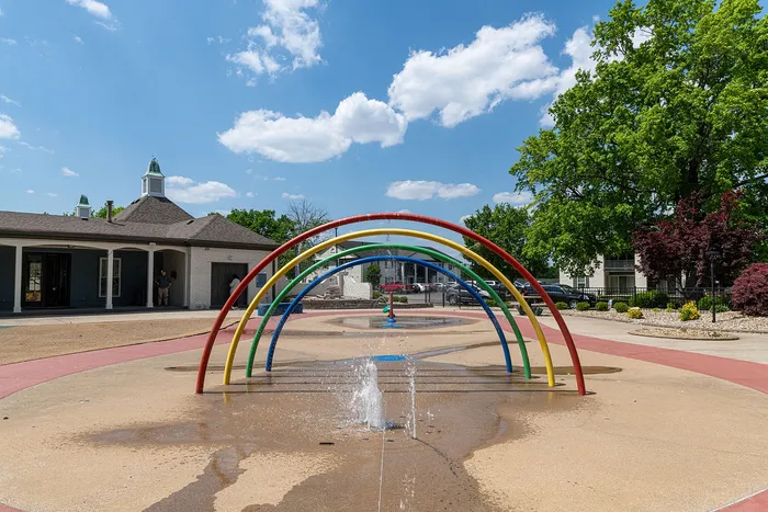 Inviting outdoor splash pad featuring vibrant rainbow arches and refreshing water fountains, set against a picturesque clubhouse and lush green surroundings.