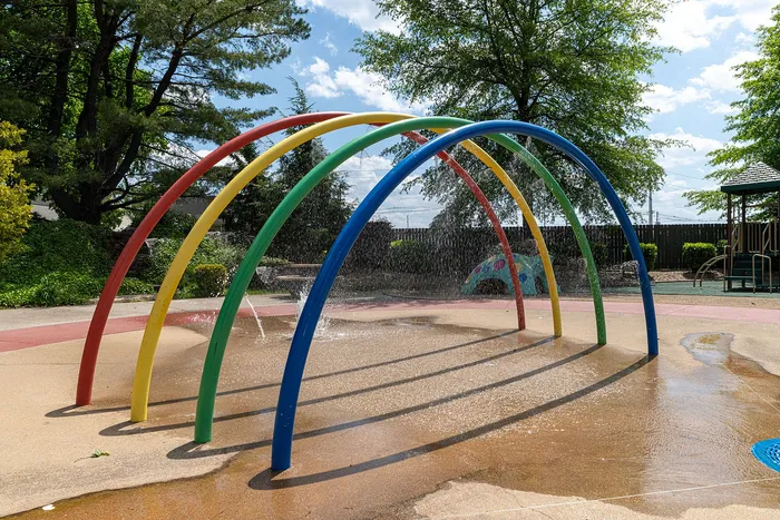 Vibrant outdoor splash pad with colorful arches and playful water features, surrounded by lush greenery and sunny blue skies.