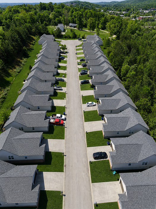 Charming aerial view of a suburban neighborhood with rows of identical houses lining a well-maintained street, surrounded by lush greenery and beautiful landscape.