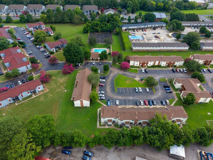 Aerial view of a charming apartment complex surrounded by lush greenery, featuring multiple buildings with red roofs, vibrant pink trees, and a refreshing community swimming pool.