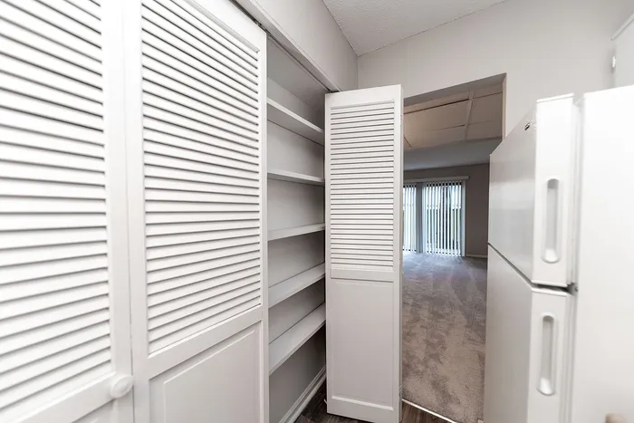 Bright apartment with open shelving, sleek white closet doors, and expansive living area with large windows and vertical blinds.