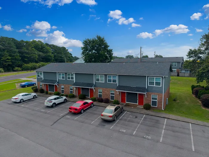 Attractive townhouse complex with modern red-brick facade, ample parking, and lush green surroundings under a vibrant blue sky.