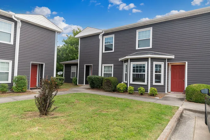 Charming modern townhouse exterior with sleek gray siding and vibrant red doors, surrounded by lush greenery and bright blue skies.