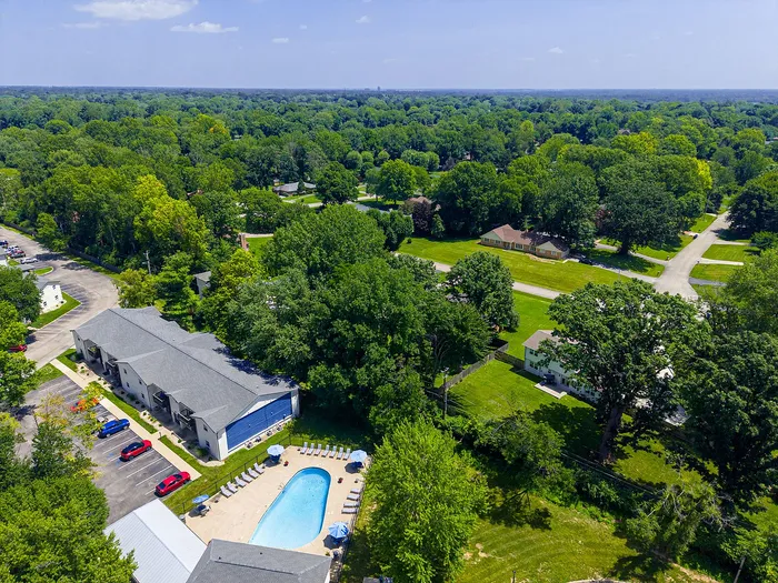 Aerial view of serene apartment community surrounded by lush green trees with a sparkling pool, ample parking, and inviting pathways.