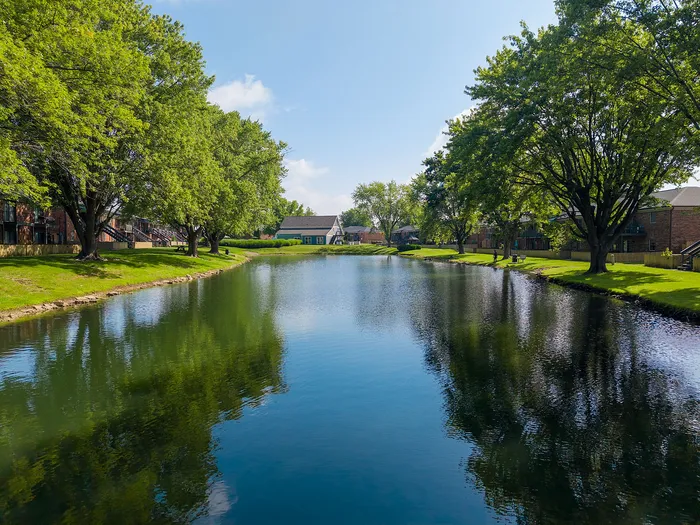 Serene residential view with lush green trees lining a tranquil pond, offering a picturesque setting for peaceful living.