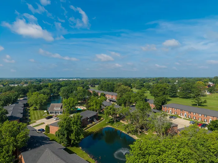 Aerial view of a serene suburban community with lush green landscapes, tranquil ponds, and charming brick buildings under a vibrant blue sky.