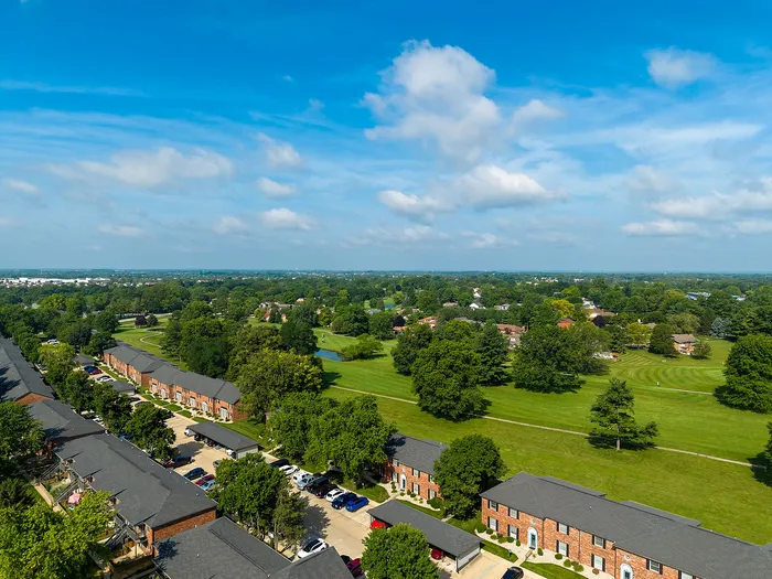 Aerial view of charming brick apartments surrounded by lush greenery and a beautiful golf course, set against a vibrant blue sky.
