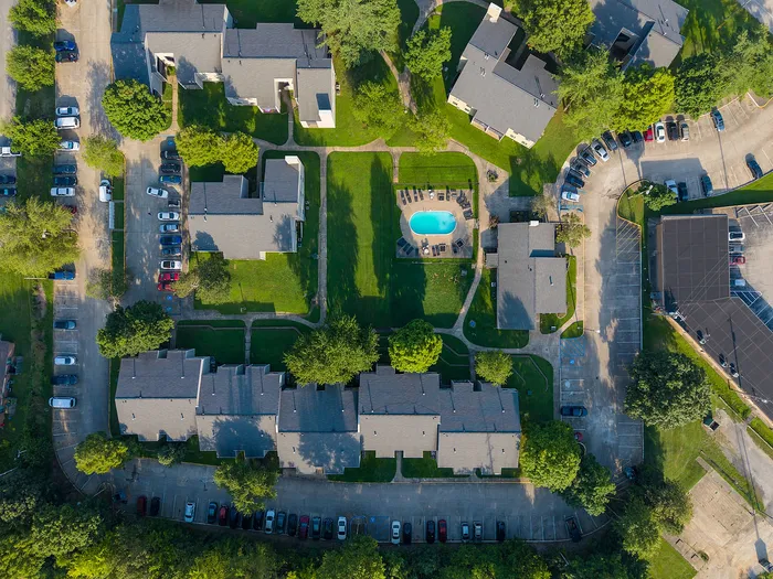 Aerial view of a charming apartment complex featuring landscaped greenery, a central swimming pool, and ample parking spaces, offering a serene living environment.