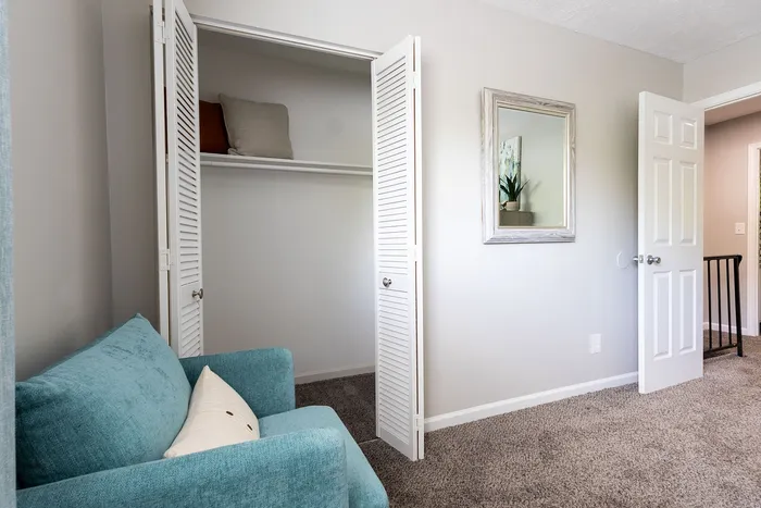 Cozy room with a plush teal chair, open white closet, and elegant wall mirror reflecting natural light.