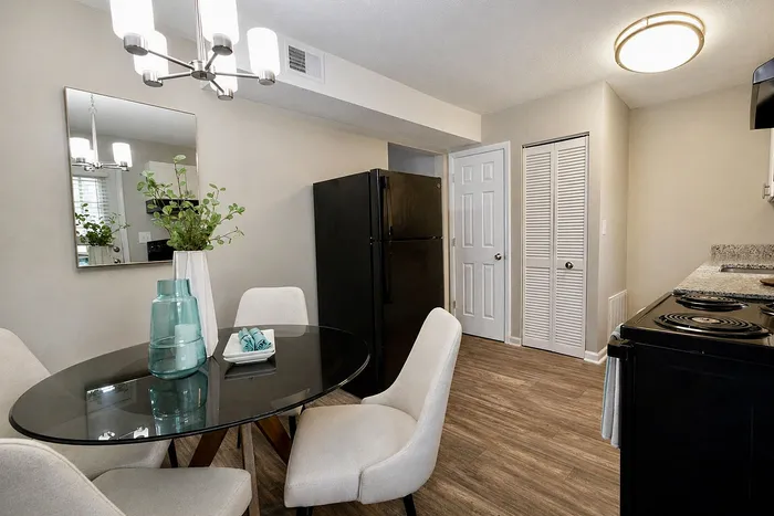 Inviting dining area with modern chandelier, elegant glass table, and cozy seating, adjacent to a sleek black fridge in a stylish kitchen with wooden flooring.